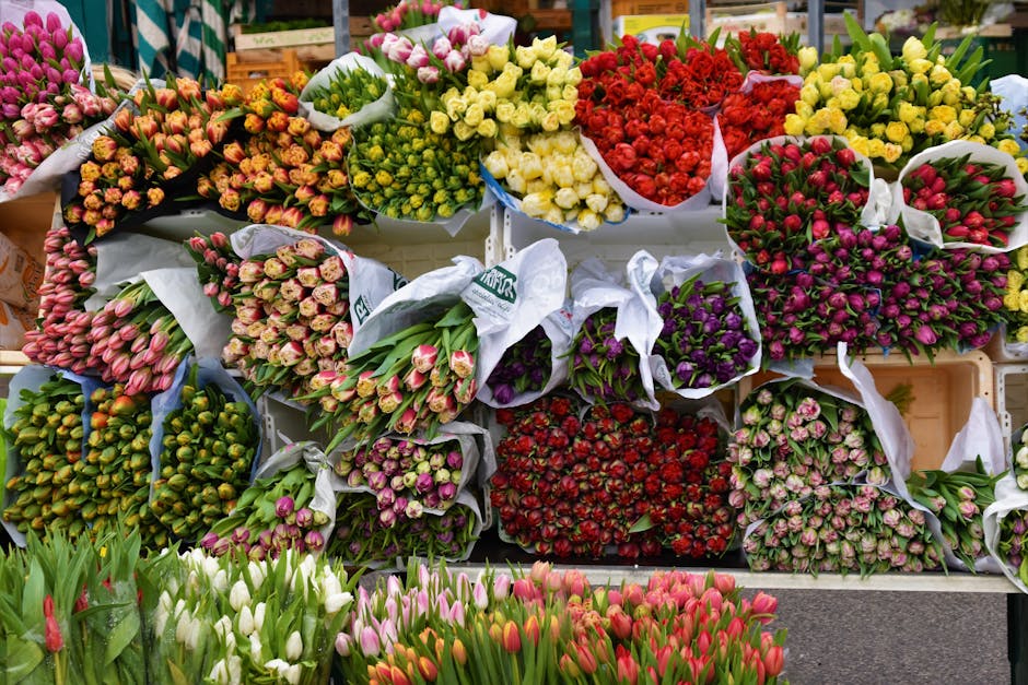 A vibrant display of various flower bouquets arranged on a market stall, featuring an assortment of tulips, roses, and ranunculus in colors including pink, red, yellow, purple, and white. The flowers